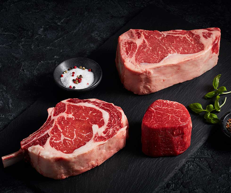 Two raw ribeye steaks and one raw filet mignon resting on a black wooden board